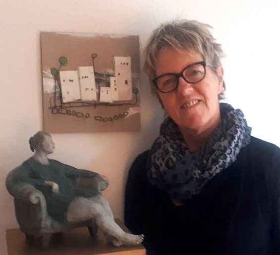Eine Frau mit kurzen, blonden Haaren steht neben einem Bild mit angedeuteten Häusern und einer getöpferten Figur von einer Frau in einem Sessel.