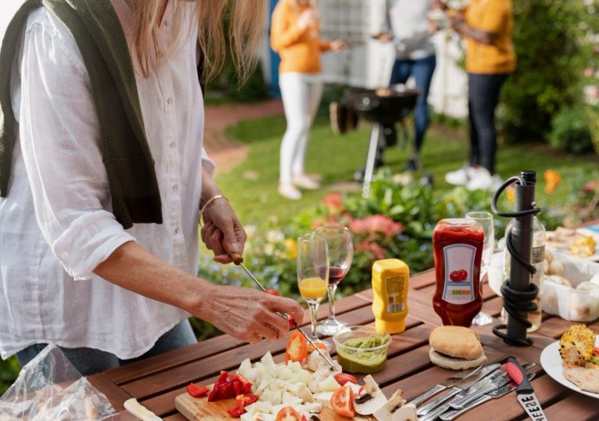Gartentisch mit verschiedenen Grillso&szlig;en, Gl&auml;sern und einem Brett mit geschnittenen Gem&uuml;se, davor eine Frau, die das Gem&uuml;se auf einen Spie&szlig; steckt. Im Hintergrund sieht man mehrere Menschen um einen Grill stehen.