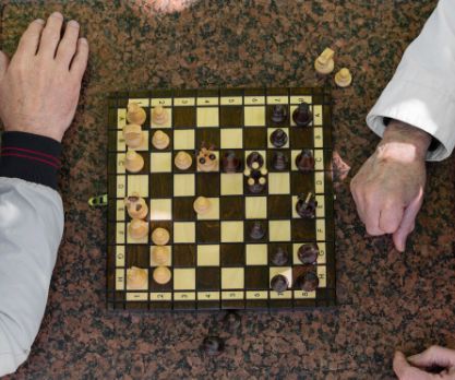 Bild von einem Schachbrett mit Schachfiguren in einem laufenden Spiel. Neben dem Schachbrett stehen auf beiden Seiten jeweils zwei Spielfiguren und man sieht die Unterarme und Hände von zwei älteren Menschen.