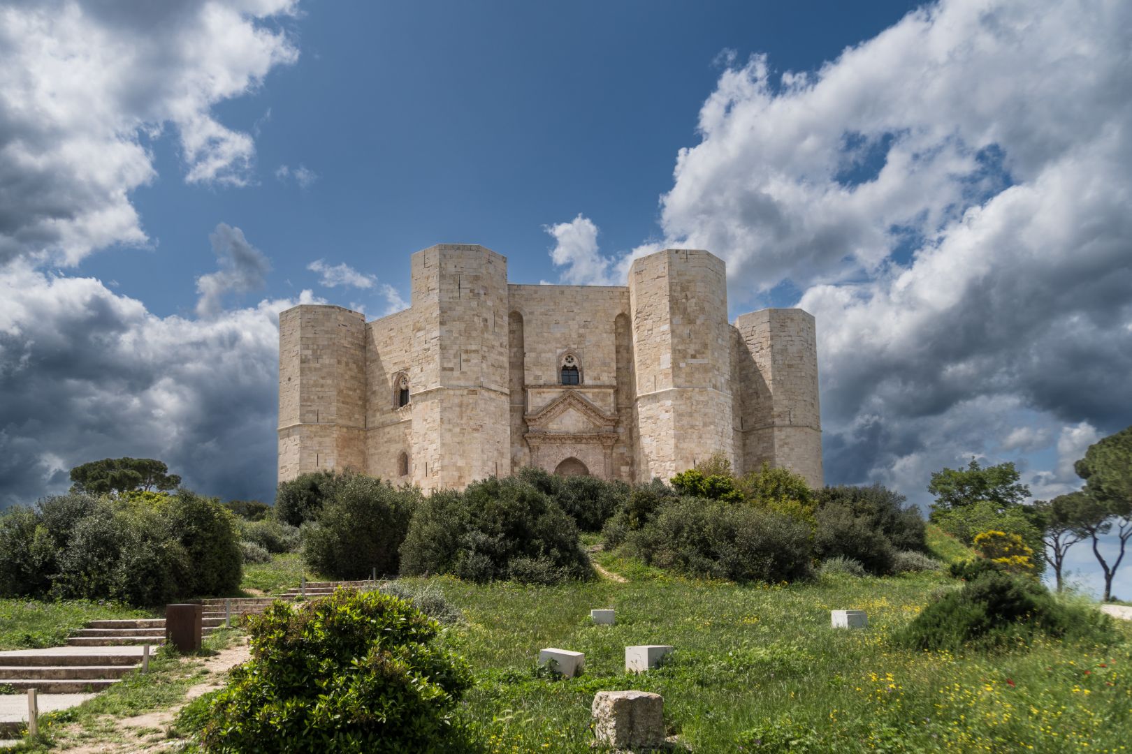 Apulien Bild des Castel del Monte - gemauerte mehreckige Burg mit vier achteckigen Türmen auf einem Hügel umgeben von Büschen, mit einer steinernen Treppe die hinauf führt